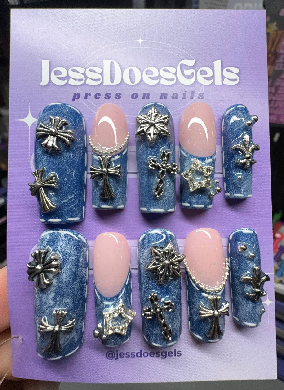 A 10-piece set of blue glitter press-on nails with silver metallic cross and snowflake embellishments, displayed on a purple card labeled 'JessDoesGels'.