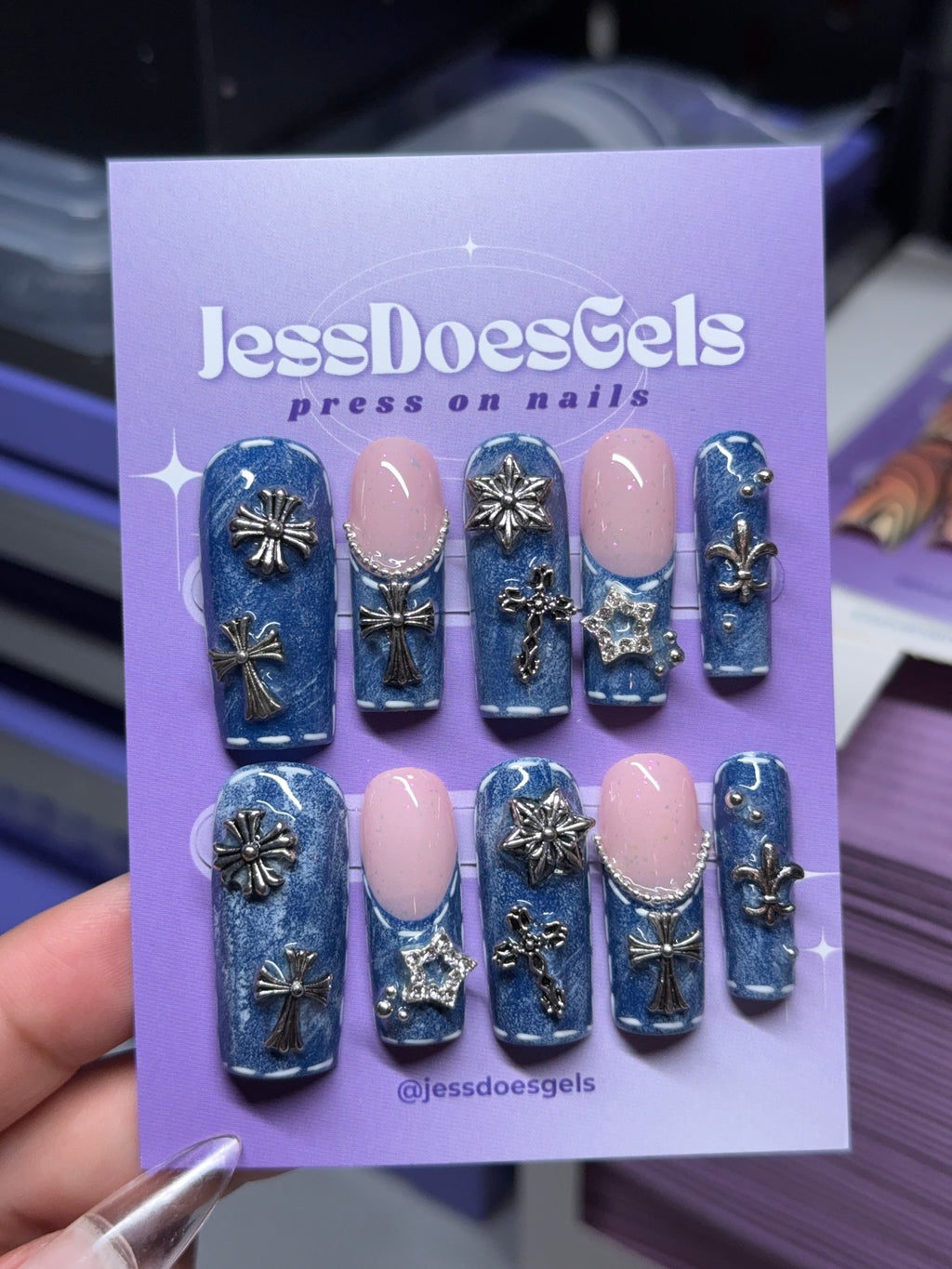 A hand holds a purple card displaying a set of press-on nails. The nails are blue with metallic silver crosses, flowers, and stars, and two pink nails.